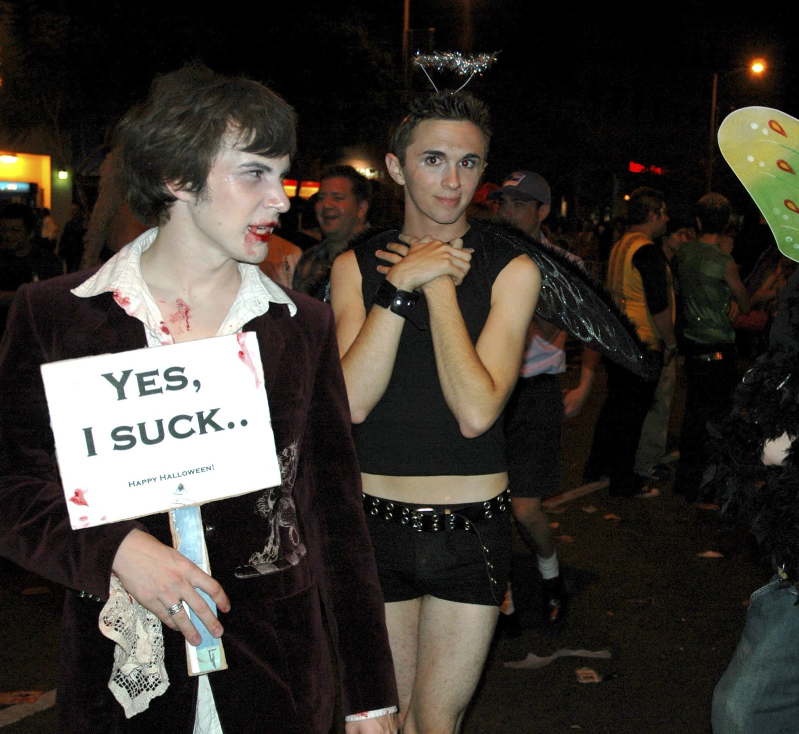 Two young men in Halloween costumes in a West Hollywood crowd, one holding a sign that reads Yes, I suck